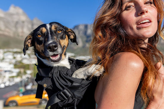 Vrouw met golvend roodbruin haar draagt een kleine hond in een zwarte draagtas op haar rug, buiten in een zonnige bergachtige omgeving met stad en kust op de achtergrond.